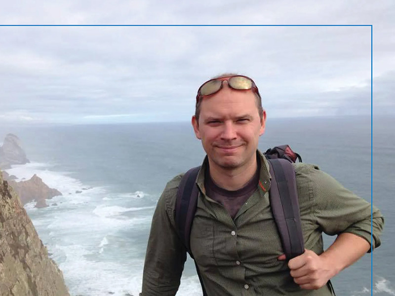 USDSer Bill Chapman wears a backpack standing at a cliff overlook with crashing waves.