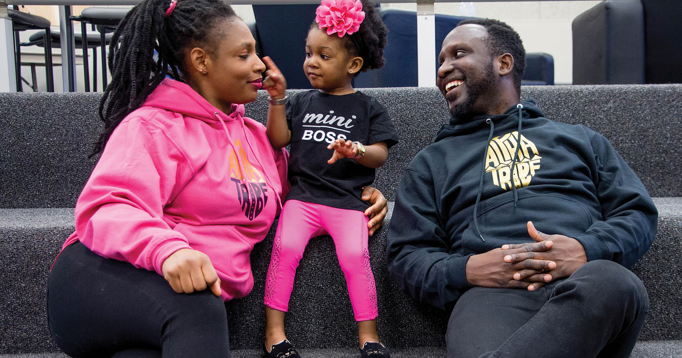 Photograph of a mother and father sitting with their young daughter. The daughter is touching their mother's nose while the father is looking on smiling.