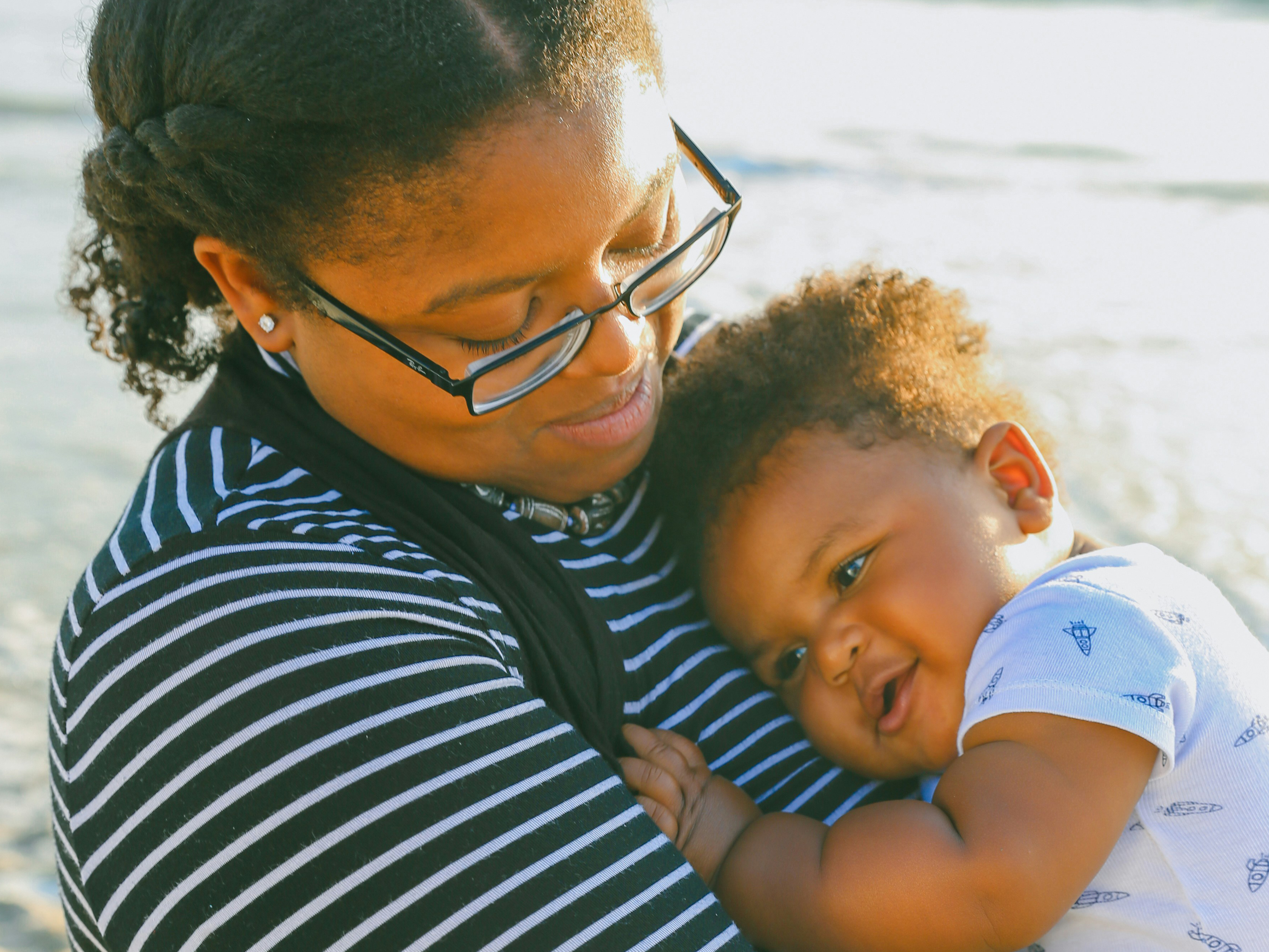 African American woman wearing glasses looks down at baby she holds against her chest.